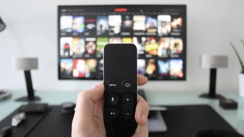 Mão segurando o controle de um aparelho de streaming diante de uma TV com catálogo de títulos.